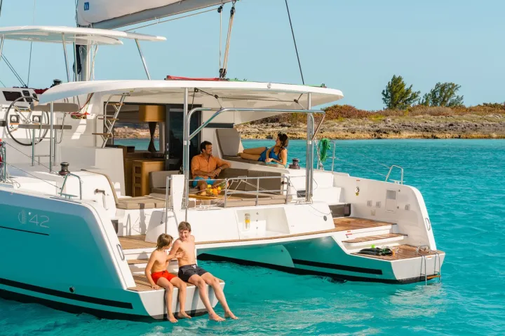 People relaxing on a yacht in turquoise water near a rocky shore under a clear sky.